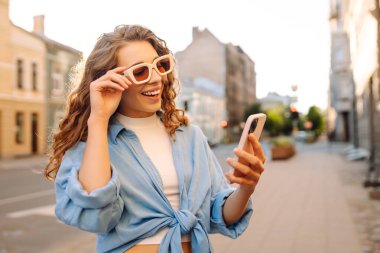 Kalabalık bir caddede telefon kullanan güneş gözlüklü genç bir kadının portresi. Neşeli bir kadın güneşli bir günün tadını çıkarıyor, şehirde geziniyor ve bloglar yazıyor. Yaşam biçimi.