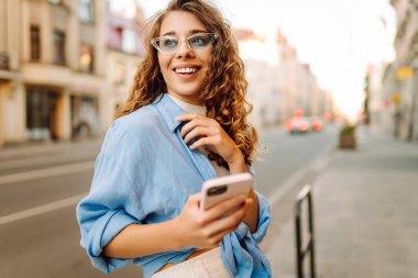 Kalabalık bir caddede telefon kullanan güneş gözlüklü genç bir kadının portresi. Neşeli bir kadın güneşli bir günün tadını çıkarıyor, şehirde geziniyor ve bloglar yazıyor. Yaşam biçimi.