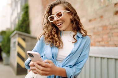 Portrait of a young woman in sunglasses using a phone on a busy city street. A cheerful woman enjoys a sunny day, strolls through the city, and blogs. Lifestyle.