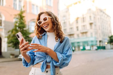 Kalabalık bir caddede telefon kullanan güneş gözlüklü genç bir kadının portresi. Neşeli bir kadın güneşli bir günün tadını çıkarıyor, şehirde geziniyor ve bloglar yazıyor. Yaşam biçimi.