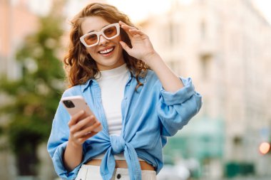 Kalabalık bir caddede telefon kullanan güneş gözlüklü genç bir kadının portresi. Neşeli bir kadın güneşli bir günün tadını çıkarıyor, şehirde geziniyor ve bloglar yazıyor. Yaşam biçimi.