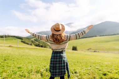 Şapkalı mutlu kadın gün batımında yeşil tepelerin tadını çıkarıyor, özgür hissediyor. Sırt çantalı bir kadın dağlarda yürüyüş parkurunda yürüyor, manzarayı seyrediyor. Özgürlük, rahatlama ve doğa kavramları..