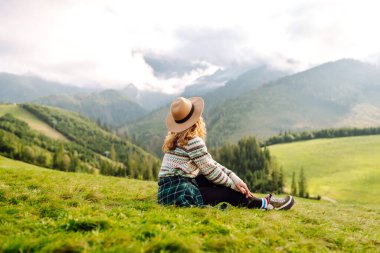 Şapkalı mutlu kadın gün batımında yeşil tepelerin tadını çıkarıyor, özgür hissediyor. Sırt çantalı bir kadın dağlarda yürüyüş parkurunda yürüyor, manzarayı seyrediyor. Özgürlük, rahatlama ve doğa kavramları..