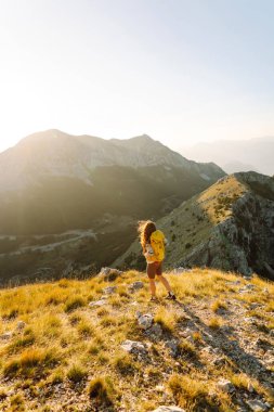 A young woman with hiking backpack stands on mountain trail on sunny day. Female hiker with a yellow backpack enjoys mountain scenery at sunset. Concepts of adventure, freedom. Active lifestyle.