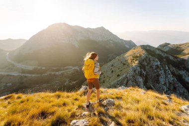 Sırt çantalı genç bir kadın güneşli bir günde dağ yolunda duruyor. Sarı sırt çantalı bir bayan yürüyüşçü gün batımında dağ manzarasının tadını çıkarıyor. Macera kavramları, özgürlük. Etkin yaşam biçimi.