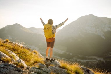 Sırt çantalı genç bir kadın güneşli bir günde dağ yolunda duruyor. Sarı sırt çantalı bir bayan yürüyüşçü gün batımında dağ manzarasının tadını çıkarıyor. Macera kavramları, özgürlük. Etkin yaşam biçimi.