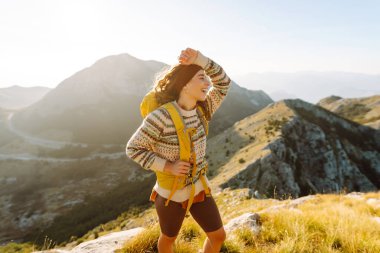 Sırt çantalı genç bir kadın güneşli bir günde dağ yolunda duruyor. Sarı sırt çantalı bir bayan yürüyüşçü gün batımında dağ manzarasının tadını çıkarıyor. Macera kavramları, özgürlük. Etkin yaşam biçimi.