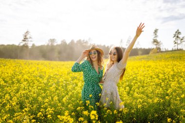 Elbiseli mutlu kız arkadaşlar güneş ışınlarındaki sarı bir tarlada eğleniyorlar. İki genç kadın geziniyor, dışarıda birlikte vakit geçiriyorlar. Arkadaşlık ve moda kavramları.