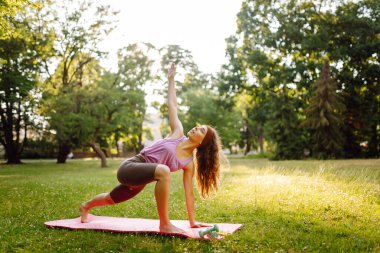 Atletik bir kadın parkta minder üzerinde yoga yapıyor. Spor elbiseli genç bir kadın gün batımında esner ya da egzersiz yapar. Spor, sağlık ve eğlence kavramı.