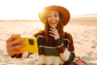 Portrait of a woman in a hat taking a selfie on the beach at sunset. A beautiful woman enjoys the seascape and blogs outdoors. Concepts: blogging, relaxation, weekends.