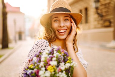 Güzel bir kadın bir buket kır çiçeğiyle güneşli bir günün tadını çıkarır. Şapkalı tatlı bir kadın bir buket çiçekle dışarıda eğleniyor. Konseptler: güzellik, moda ve eğlence.