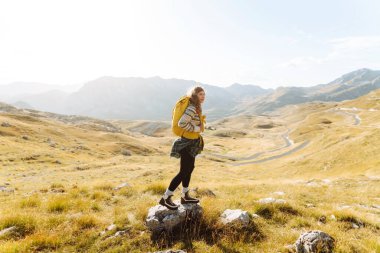 Sarı sırt çantalı mutlu bir yürüyüşçü yürüyüş parkurunda dağ manzarasının tadını çıkarıyor. Genç kadın açık havanın özgürlüğünü hissediyor. Yürüyüş, özgürlük ve macera kavramı.