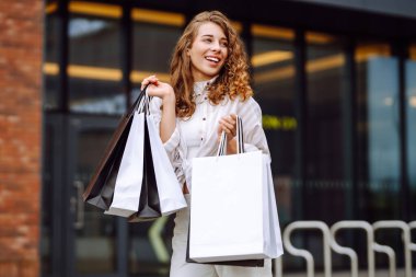 Bir mağazanın önünde elinde poşetlerle gülümseyen genç bir kadın. O, kalabalık bir alışveriş alanında büyük vitrinlerin önünde duruyor, bir alışveriş gezisinden sonra memnun görünüyor.