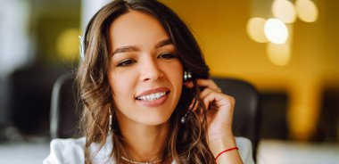 Kulaklık takan bir kadın telefonda konuşurken gülümsüyor. Masasında oturuyor, parlak bir ofis alanında bilgisayarının başında çalışıyor. İşiyle meşgul görünüyor ve görevlerini tamamlamaya odaklanmış..