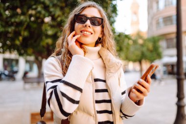 Şehir meydanında telefonu olan şık bir kadının portresi. Güzel bir kadın güneşli bir günde gezinti yapmaktan ve telefonunda blog yazmaktan hoşlanır. Yaşam tarzı. Turizm konsepti.
