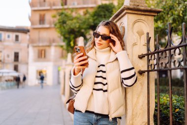 Şehir meydanında telefonu olan şık bir kadının portresi. Güzel bir kadın güneşli bir günde gezinti yapmaktan ve telefonunda blog yazmaktan hoşlanır. Yaşam tarzı. Turizm konsepti.