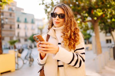 Şehir meydanında telefonu olan şık bir kadının portresi. Güzel bir kadın güneşli bir günde gezinti yapmaktan ve telefonunda blog yazmaktan hoşlanır. Yaşam tarzı. Turizm konsepti.