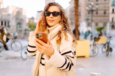 Şehir meydanında telefonu olan şık bir kadının portresi. Güzel bir kadın güneşli bir günde gezinti yapmaktan ve telefonunda blog yazmaktan hoşlanır. Yaşam tarzı. Turizm konsepti.