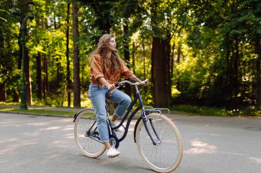 Güneşli bir parkta bisiklet süren genç bir kadın. Neşeli bir turist dışarıda bisiklet sürüyor, doğanın tadını çıkarıyor. Konsept: ulaşım, boş zaman, aktif yaşam tarzı.