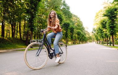 Güneşli bir parkta bisiklet süren genç bir kadın. Neşeli bir turist dışarıda bisiklet sürüyor, doğanın tadını çıkarıyor. Konsept: ulaşım, boş zaman, aktif yaşam tarzı.