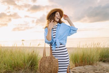 Hasır şapkalı neşeli bir kadın kumlu bir sahilde çanta taşıyor. Güzel bir kadın gün batımını ve deniz manzarasını sever. Tatil, hafta sonu ve moda kavramı.