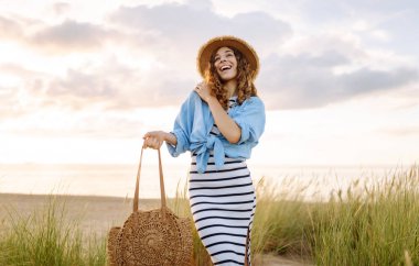 Hasır şapkalı neşeli bir kadın kumlu bir sahilde çanta taşıyor. Güzel bir kadın gün batımını ve deniz manzarasını sever. Tatil, hafta sonu ve moda kavramı.