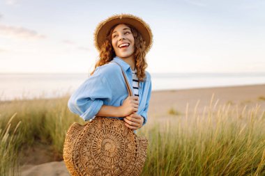 Hasır şapkalı neşeli bir kadın kumlu bir sahilde çanta taşıyor. Güzel bir kadın gün batımını ve deniz manzarasını sever. Tatil, hafta sonu ve moda kavramı.