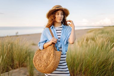 Hasır şapkalı neşeli bir kadın kumlu bir sahilde çanta taşıyor. Güzel bir kadın gün batımını ve deniz manzarasını sever. Tatil, hafta sonu ve moda kavramı.