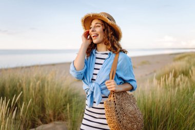 Hasır şapkalı neşeli bir kadın kumlu bir sahilde çanta taşıyor. Güzel bir kadın gün batımını ve deniz manzarasını sever. Tatil, hafta sonu ve moda kavramı.