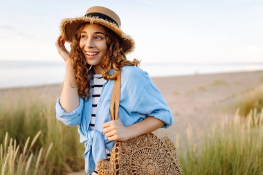 Hasır şapkalı neşeli bir kadın kumlu bir sahilde çanta taşıyor. Güzel bir kadın gün batımını ve deniz manzarasını sever. Tatil, hafta sonu ve moda kavramı.
