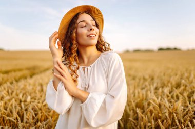 Şapkalı tatlı bir kadın gün batımında buğday tarlasında duruyor. Güzel bir kadın gün batımından zevk alır ve bir çiftlik arazisinde gezinir. Güzellik kavramı, doğa.