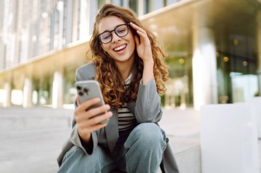 Gözlük takan neşeli bir kadının portresi ve modern bir ofis binasının arka planında telefon kullanmak. Şık bir kadın blogu yapıyor ve güneşli bir gün geçiriyor..