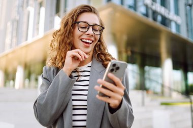 Gözlük takan neşeli bir kadının portresi ve modern bir ofis binasının arka planında telefon kullanmak. Şık bir kadın blogu yapıyor ve güneşli bir gün geçiriyor..