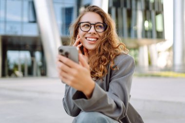Gözlük takan neşeli bir kadının portresi ve modern bir ofis binasının arka planında telefon kullanmak. Şık bir kadın blogu yapıyor ve güneşli bir gün geçiriyor..