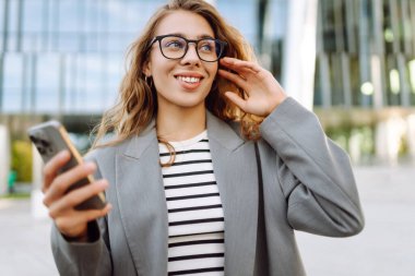 Gözlük takan neşeli bir kadının portresi ve modern bir ofis binasının arka planında telefon kullanmak. Şık bir kadın blogu yapıyor ve güneşli bir gün geçiriyor..