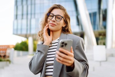 Gözlük takan neşeli bir kadının portresi ve modern bir ofis binasının arka planında telefon kullanmak. Şık bir kadın blogu yapıyor ve güneşli bir gün geçiriyor..