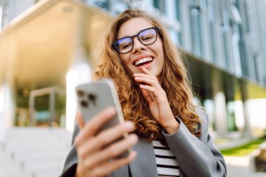 Gözlük takan neşeli bir kadının portresi ve modern bir ofis binasının arka planında telefon kullanmak. Şık bir kadın blogu yapıyor ve güneşli bir gün geçiriyor..