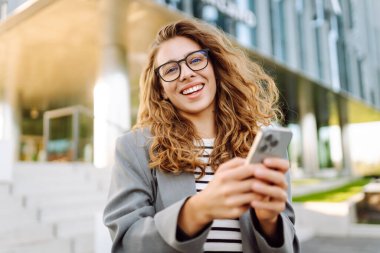 Gözlük takan neşeli bir kadının portresi ve modern bir ofis binasının arka planında telefon kullanmak. Şık bir kadın blogu yapıyor ve güneşli bir gün geçiriyor..