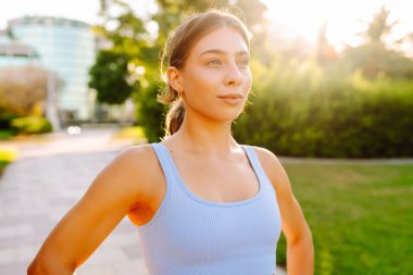 Güneşli bir parkta kollarını açmış gülümseyen bir kadın. Spor giyim sektöründe güzel bir kadın antrenmandan önceki güneşli günün tadını çıkarır. Spor konsepti, açık hava..