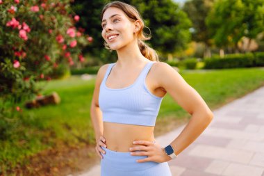 Güneşli bir parkta kollarını açmış gülümseyen bir kadın. Spor giyim sektöründe güzel bir kadın antrenmandan önceki güneşli günün tadını çıkarır. Spor konsepti, açık hava..
