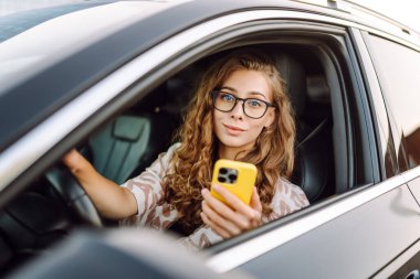 Araba kullanırken telefonla konuşan gözlüklü genç bir kadının portresi. Güneşli bir günde modern bir arabada elinde telefon tutan bir kadın sürücü. Ulaşım ve teknoloji kavramı.