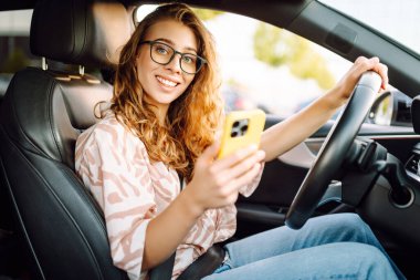 Araba kullanırken telefonla konuşan gözlüklü genç bir kadının portresi. Güneşli bir günde modern bir arabada elinde telefon tutan bir kadın sürücü. Ulaşım ve teknoloji kavramı.