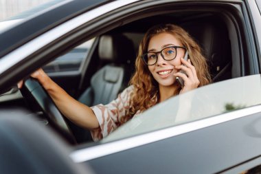Araba kullanırken telefonla konuşan gözlüklü genç bir kadının portresi. Güneşli bir günde modern bir arabada elinde telefon tutan bir kadın sürücü. Ulaşım ve teknoloji kavramı.