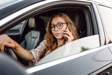 Araba kullanırken telefonla konuşan gözlüklü genç bir kadının portresi. Güneşli bir günde modern bir arabada elinde telefon tutan bir kadın sürücü. Ulaşım ve teknoloji kavramı.