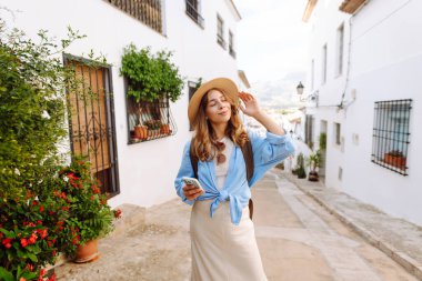 Şapkalı ve telefonlu güzel bir turist şehrin sokaklarında güneşli bir günün tadını çıkarıyor. Sırt çantası blogları olan ve gün batımında gezen neşeli bir kadın. Turizm konsepti, hafta sonu. Etkin yaşam biçimi.