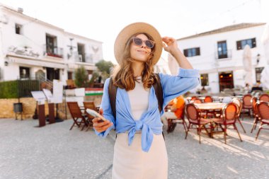 Şapkalı ve telefonlu güzel bir turist şehrin sokaklarında güneşli bir günün tadını çıkarıyor. Sırt çantası blogları olan ve gün batımında gezen neşeli bir kadın. Turizm konsepti, hafta sonu. Etkin yaşam biçimi.