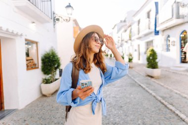 Şapkalı ve telefonlu güzel bir turist şehrin sokaklarında güneşli bir günün tadını çıkarıyor. Sırt çantası blogları olan ve gün batımında gezen neşeli bir kadın. Turizm konsepti, hafta sonu. Etkin yaşam biçimi.