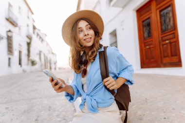 Şapkalı ve telefonlu güzel bir turist şehrin sokaklarında güneşli bir günün tadını çıkarıyor. Sırt çantası blogları olan ve gün batımında gezen neşeli bir kadın. Turizm konsepti, hafta sonu. Etkin yaşam biçimi.