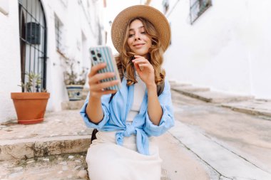 Şapkalı ve telefonlu güzel bir turist şehrin sokaklarında güneşli bir günün tadını çıkarıyor. Sırt çantası blogları olan ve gün batımında gezen neşeli bir kadın. Turizm konsepti, hafta sonu. Etkin yaşam biçimi.