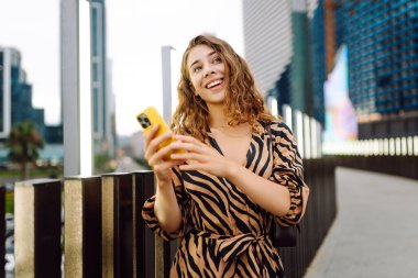 Elinde telefon olan genç bir kadın, şehir merkezine bakan yaya köprüsünde duruyor. Bir turist yürüyüş yapmaktan, mesajlaşmaktan ya da dışarıda blog yazmaktan hoşlanır. Günlük konsepti, yürüyüş.